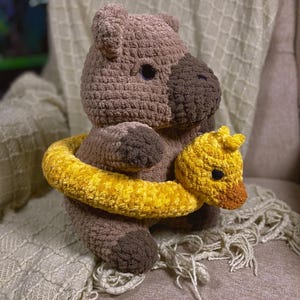 May include: A brown crocheted capybara plush toy with a dark brown snout and black eyes. The capybara is wearing a bright yellow crocheted duck-shaped pool float around its middle, featuring a small duck head with a brown beak and black eyes.