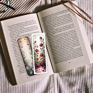 May include: Two decorative bookmarks with floral designs. One features a watercolor style with red roses and a reflection, the other with a light background and delicate flowers. The bookmarks are placed on an open book.
