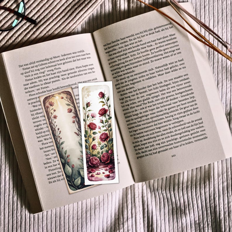 May include: Two decorative bookmarks with floral designs. One features a watercolor style with red roses and a reflection, the other with a light background and delicate flowers. The bookmarks are placed on an open book.