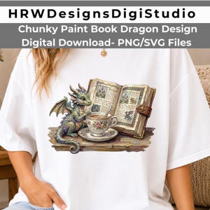 以下が含まれることがあります： 白いTシャツに、開いたイラスト入りの本と花柄のティーカップ、ソーサーの横に座る小さな緑色のドラゴンのグラフィックプリントが施されています。デザインには、城やドラゴンのモチーフが詳細に描かれています。