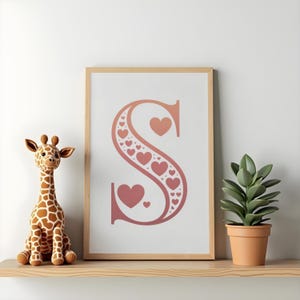 Op de afbeelding: Een ingelijste print met de letter "S" in roze tinten, versierd met hartmotieven. Een knuffelgiraf en een plant in een pot staan op een houten plank, wat een decoratie vormt.