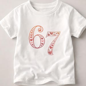 Op de afbeelding: Wit T-shirt met het nummer 67 in een rood-oranje verloop, gevuld met hartvormen. Het shirt heeft korte mouwen en een ronde hals.