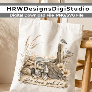 May include: A white canvas tote bag featuring a printed illustration of a sea turtle wearing a striped bow, resting on a beach with seashells, tall grass, two seagulls perched on driftwood, and a sunset, with the text "slow down, soak it in".