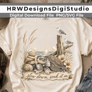 May include: A cream-colored t-shirt featuring a graphic of a sea turtle wearing a striped bow, stacked seashells, tall beach grass, two seagulls perched on driftwood, and a sunrise over the ocean with the text, "slow down, soak it in."