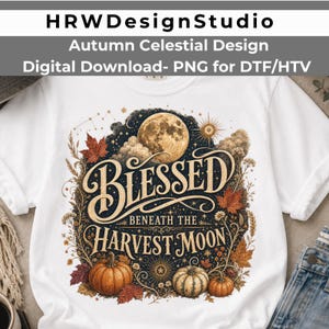 May include: A white t-shirt featuring a graphic print with the text BLESSED BENEATH THE HARVEST MOON. The design includes a large full moon, autumn leaves, pumpkins, stars, and clouds in shades of orange, brown, gold, and dark blue.