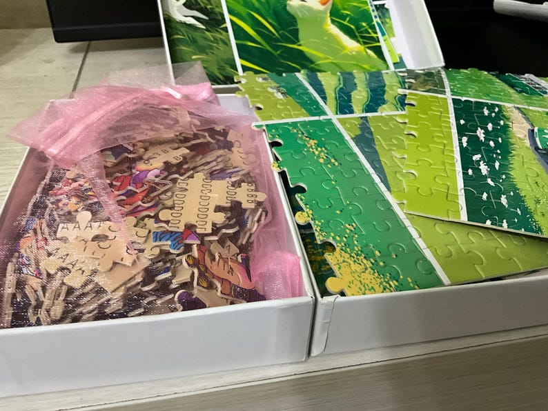 Pu&ograve; includere: Una scatola bianca aperta contenente un puzzle con pezzi verdi e marroni. La scatola rivela i pezzi del puzzle e un sacchetto di rete rosa. Il puzzle raffigura una rana in un'area erbosa.