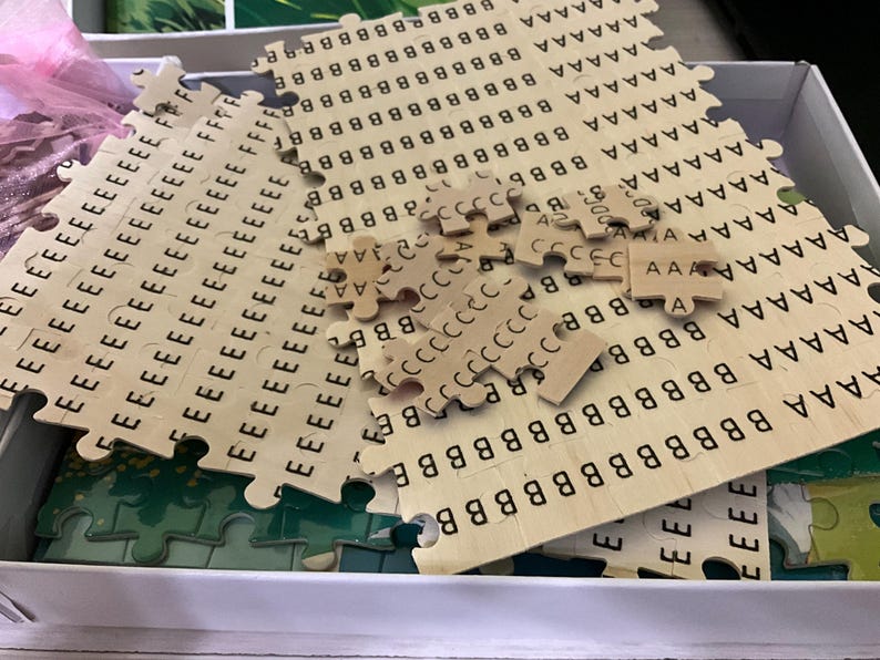Pu&ograve; includere: Un puzzle di lettere in legno parzialmente assemblato con pezzi ad incastro. I pezzi del puzzle sono marrone chiaro con lettere nere stampate. Alcuni pezzi sono sparsi, altri sono ancora nella scatola. Le lettere includono A, B, C ed E.