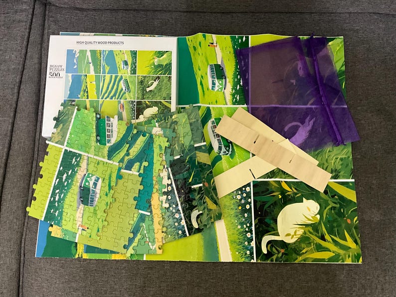 Pu&ograve; includere: Un puzzle da 500 pezzi parzialmente assemblato con una scena di paesaggio verde e bianco. I pezzi del puzzle sono sparsi su una base stampata colorata, con un sacchetto viola e supporti in legno. La scatola recita "HIGH QUALITY WOOD PRODUCTS".