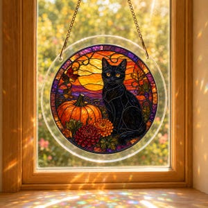 Puede incluir: Un atrapasueños circular de vidrieras que muestra un gato negro sentado junto a una gran calabaza naranja y flores otoñales coloridas. El fondo presenta una luna llena amarilla vibrante y un cielo púrpura, suspendido por una fina cadena de metal.