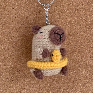 以下が含まれることがあります： ベージュの羊と茶色のアクセントが特徴の手作りのかぎ針編みのキーホルダー。黄色いひよこを抱えています。羊は茶色の顔と耳をしており、ひよこは黄色のバンドで囲まれています。キーホルダーにはシルバーのリングと青いビーズが付いています。
