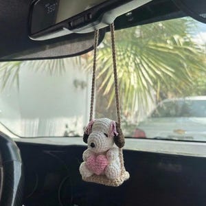 Puede incluir: Amuleto de coche de ganchillo con un perro blanco con orejas rosas y cabeza marr&oacute;n, sentado en un columpio. El perro sostiene un coraz&oacute;n rosa. El amuleto est&aacute; suspendido por cuerdas beige, dise&ntilde;ado para colgar del espejo retrovisor de un coche.