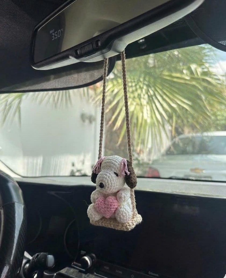 Puede incluir: Amuleto de coche de ganchillo con un perro blanco con orejas rosas y cabeza marr&oacute;n, sentado en un columpio. El perro sostiene un coraz&oacute;n rosa. El amuleto est&aacute; suspendido por cuerdas beige, dise&ntilde;ado para colgar del espejo retrovisor de un coche.