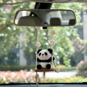 Könnte beinhalten: Ein gehäkelter Panda-Bär als Anhänger, der auf einer kleinen braunen Schaukel sitzt, die an zwei weißen Seilen hängt. Die Seile sind mit kleinen, pastellfarbenen gehäkelten Blumen in Rosa, Gelb und Weiß sowie grünen Ranken verziert.