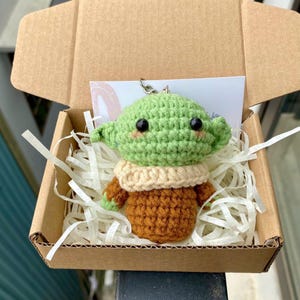 May include: A handmade crochet figure of a character with large ears, green head, and brown clothing. The figure is inside a cardboard box with paper filler. The figure has black eyes and a small smile. A small metal keychain is attached to the figure.