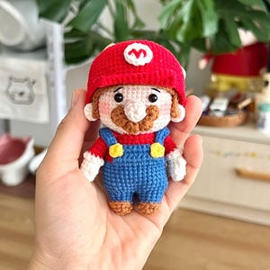 May include: Crocheted Mario doll wearing a red hat with a white 'M' symbol, a red shirt, and blue overalls. The doll has a brown mustache and is holding a hand.