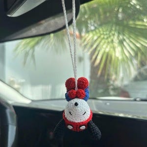 Puede incluir: Un adorno de coche de Spider-Man hecho a mano con ganchillo. El adorno est&aacute; suspendido por un hilo blanco y presenta una cabeza blanca, un cuerpo rojo y azul y brazos negros. El adorno est&aacute; colgado boca abajo.