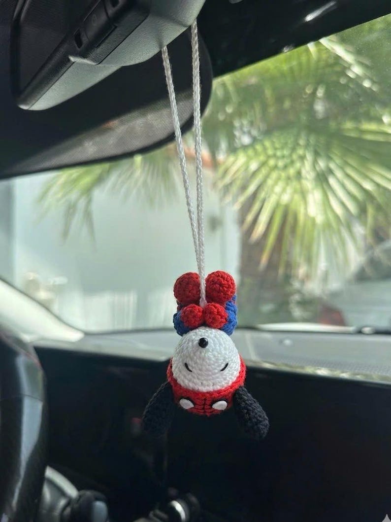 Puede incluir: Un adorno de coche de Spider-Man hecho a mano con ganchillo. El adorno est&aacute; suspendido por un hilo blanco y presenta una cabeza blanca, un cuerpo rojo y azul y brazos negros. El adorno est&aacute; colgado boca abajo.