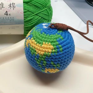 Puede incluir: Un globo terráqueo tejido a ganchillo con océanos azules, continentes verdes y masas terrestres amarillas. Una cuerda de cuero marrón está unida a la parte superior. Una madeja de hilo verde y una etiqueta con texto chino están en el fondo.