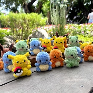May include: A collection of small, handmade crochet figures representing various creatures, including yellow characters with red cheeks, orange figures with cream bellies, and light blue characters, some holding small red and white spherical items.