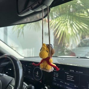 Puede incluir: Un personaje amarillo tejido a ganchillo con una bufanda roja y una gorra de aviador marr&oacute;n cuelga del espejo retrovisor de un coche. La decoraci&oacute;n hecha a mano est&aacute; suspendida por cuerdas marrones. El salpicadero y el volante del coche son visibles en el fondo.