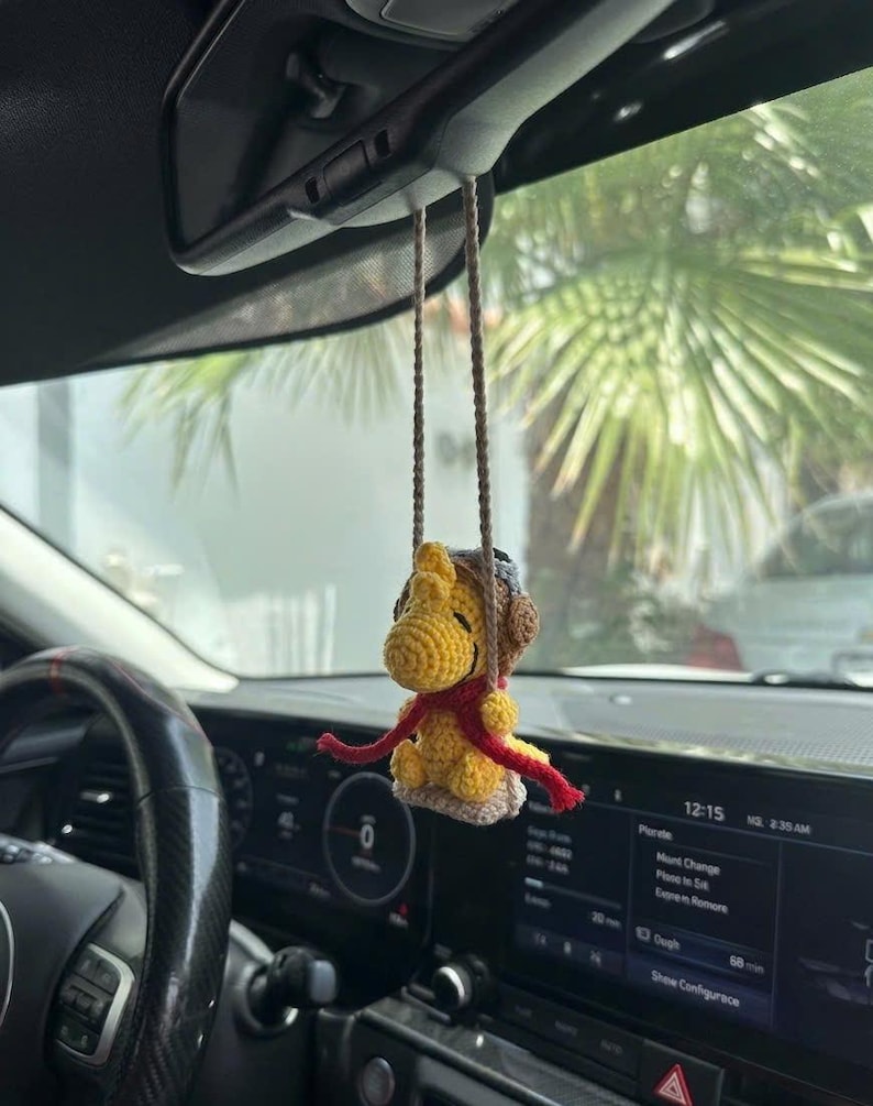 Puede incluir: Un personaje amarillo tejido a ganchillo con una bufanda roja y una gorra de aviador marr&oacute;n cuelga del espejo retrovisor de un coche. La decoraci&oacute;n hecha a mano est&aacute; suspendida por cuerdas marrones. El salpicadero y el volante del coche son visibles en el fondo.