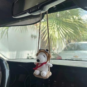 Puede incluir: Un adorno de coche de Snoopy blanco tejido a ganchillo, con un gorro de aviador marr&oacute;n y una bufanda roja, sentado en un columpio beige. El adorno cuelga de una cuerda marr&oacute;n, suspendido del espejo retrovisor de un coche.