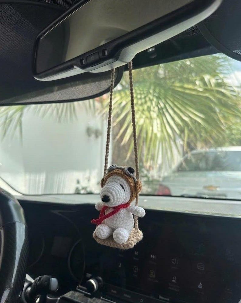 Puede incluir: Un adorno de coche de Snoopy blanco tejido a ganchillo, con un gorro de aviador marr&oacute;n y una bufanda roja, sentado en un columpio beige. El adorno cuelga de una cuerda marr&oacute;n, suspendido del espejo retrovisor de un coche.