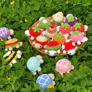 May include: A collection of colorful, crocheted turtle figurines, each designed to resemble different fruits and foods. The turtles are displayed on a wooden plate and amidst green foliage. The turtles include designs like strawberries, watermelons, and mushrooms.