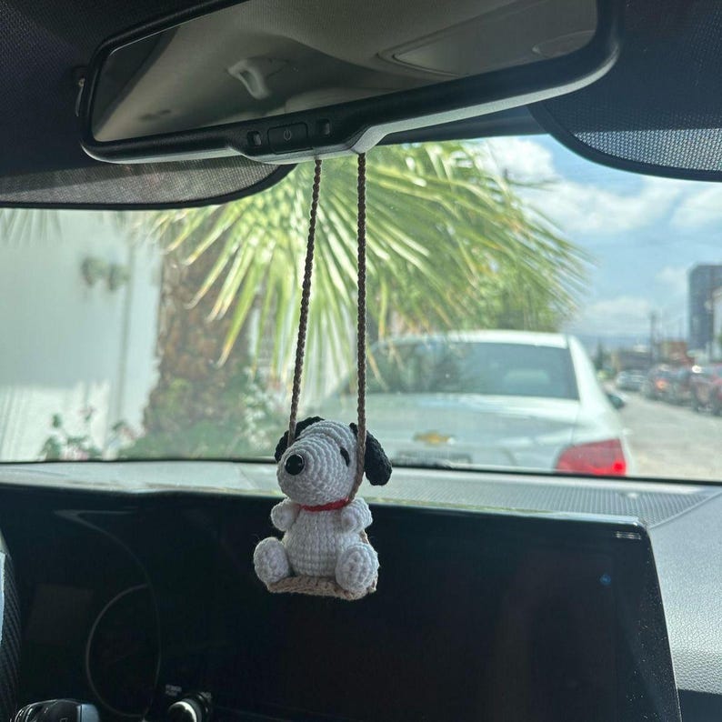 Puede incluir: Un adorno de coche de Snoopy tejido en blanco y negro con un collar rojo cuelga de una cuerda marr&oacute;n, suspendido del espejo retrovisor de un coche. El adorno tiene la forma de Snoopy sentado en una peque&ntilde;a plataforma tejida. El fondo muestra una escena soleada al aire libre.