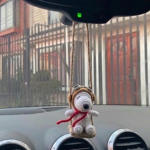 Puede incluir: Un adorno de coche de Snoopy hecho a mano. El Snoopy blanco de ganchillo lleva un gorro de aviador marr&oacute;n y una bufanda roja. El adorno cuelga de una cuerda marr&oacute;n, suspendida del espejo retrovisor del coche.
