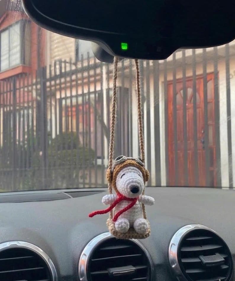 Puede incluir: Un adorno de coche de Snoopy hecho a mano. El Snoopy blanco de ganchillo lleva un gorro de aviador marr&oacute;n y una bufanda roja. El adorno cuelga de una cuerda marr&oacute;n, suspendida del espejo retrovisor del coche.