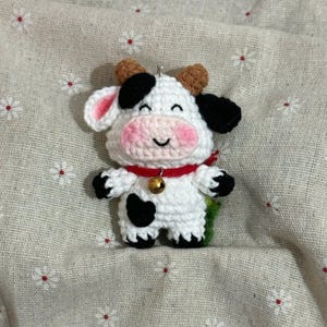 May include: A handmade crochet cow figurine. The cow is white with black spots, black ears, and brown horns. It has a pink muzzle, a red collar with a small gold bell, and a smiling face. The cow is a cute, whimsical design.