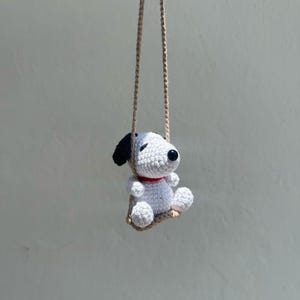 Puede incluir: Una mu&ntilde;eca Snoopy de ganchillo con orejas negras, un collar rojo y ojos negros, suspendida de un columpio de cuerda beige. El perro blanco est&aacute; sentado en un peque&ntilde;o columpio beige, creando un encantador art&iacute;culo decorativo hecho a mano.