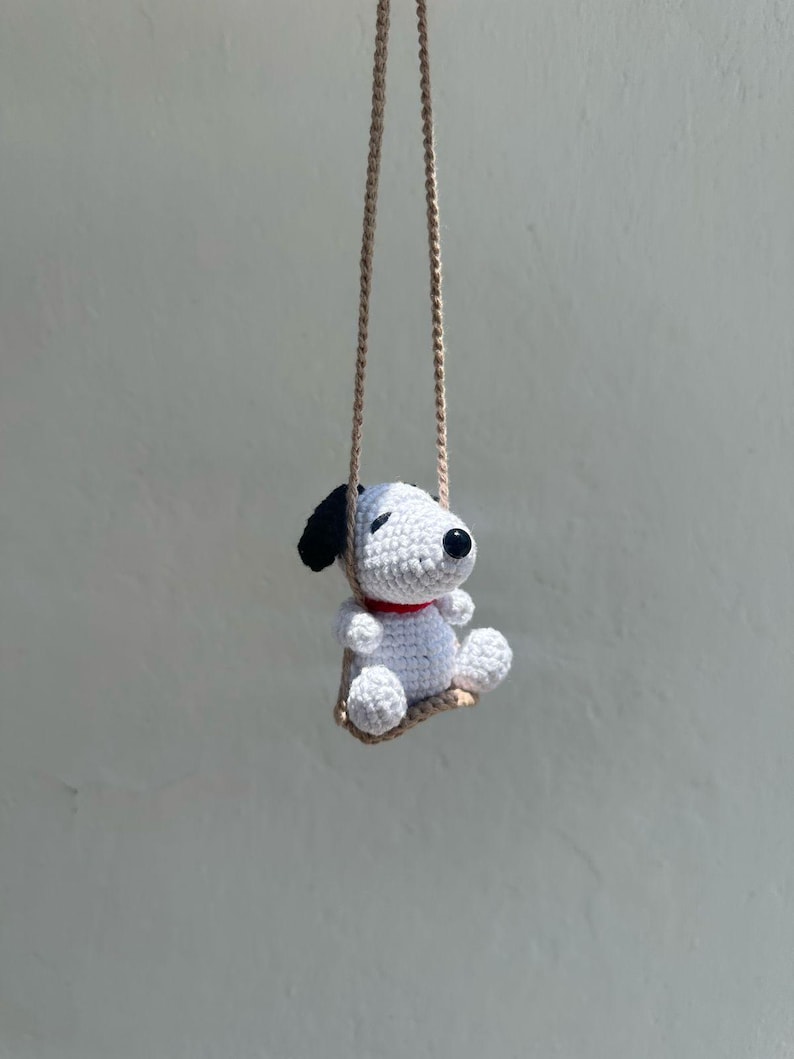 Puede incluir: Una mu&ntilde;eca Snoopy de ganchillo con orejas negras, un collar rojo y ojos negros, suspendida de un columpio de cuerda beige. El perro blanco est&aacute; sentado en un peque&ntilde;o columpio beige, creando un encantador art&iacute;culo decorativo hecho a mano.