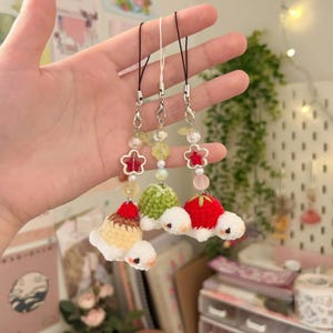 May include: Three handmade crochet charms with food-themed designs. Each charm features a small, round crocheted body in different colors, topped with a red cherry or strawberry. The charms are attached to a silver-colored clasp with a beaded chain.