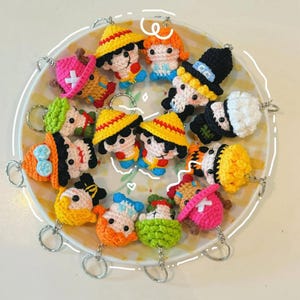 May include: A selection of crocheted character keyrings in various colours. The keyrings depict cartoon characters with unique features, such as hats, hair, and facial expressions. Each keyring has a silver ring for attachment. The keyrings are arranged on a yellow and white plate.