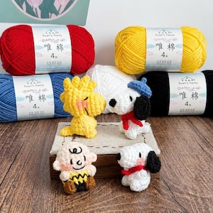 Puede incluir: Cuatro pequeñas figuras de ganchillo que representan personajes de dibujos animados, incluidos dos perros blancos con orejas negras, un pájaro amarillo y una figura humana con una camisa en zigzag, frente a cuatro ovillos de lana roja, amarilla, azul y negra.