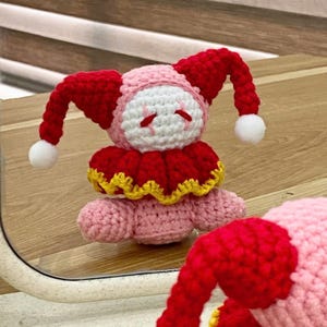 Könnte beinhalten: Handgemachte gehäkelte Narrenpuppe. Die Puppe ist überwiegend rot und rosa, mit weißen Akzenten. Sie hat einen roten Hut mit weißen Bommeln, einen roten Kragen mit gelbem Besatz und einen rosa Körper. Die Puppe steht auf einer Holzoberfläche.