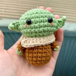 May include: A handmade crochet figure of a character with large ears and black eyes. The figure is primarily green, with a tan collar and brown body. The figure is approximately 3 inches tall.