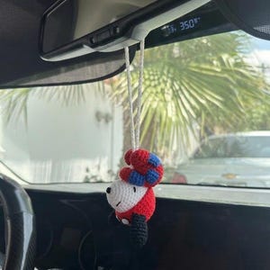 Puede incluir: Un adorno de coche hecho a mano, tejido a ganchillo con la forma de un personaje de dibujos animados. El adorno presenta una cara blanca, un cuerpo rojo, piernas negras y un sombrero rojo y azul. Cuelga del espejo retrovisor con un cord&oacute;n blanco.