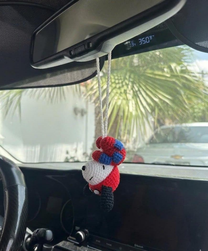 Puede incluir: Un adorno de coche hecho a mano, tejido a ganchillo con la forma de un personaje de dibujos animados. El adorno presenta una cara blanca, un cuerpo rojo, piernas negras y un sombrero rojo y azul. Cuelga del espejo retrovisor con un cord&oacute;n blanco.