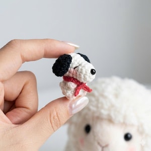 Puede incluir: Una peque&ntilde;a figura de perro de ganchillo con orejas negras, nariz negra y bufanda roja. El perro es blanco con mejillas rosadas y ojos cerrados, sostenido entre los dedos. El fondo est&aacute; borroso, con un objeto blanco y esponjoso.