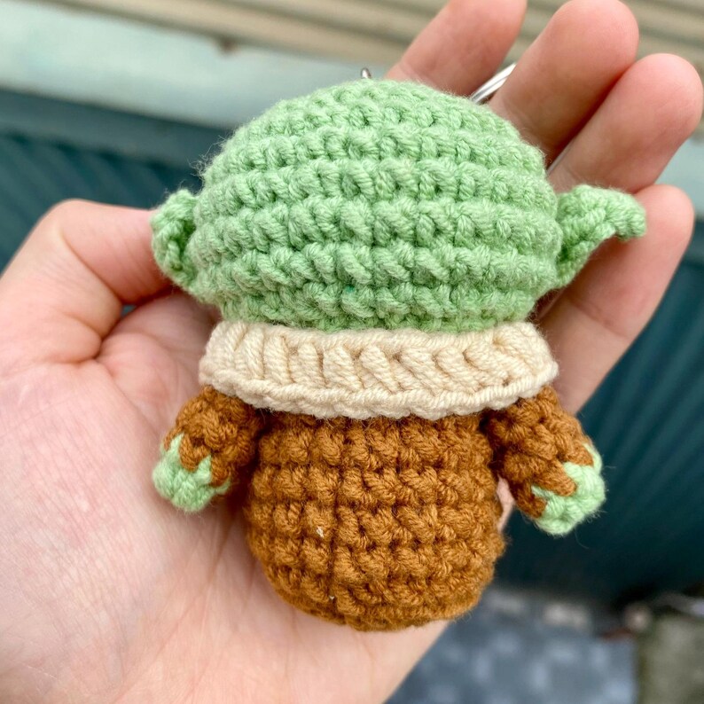 May include: A handmade crochet keychain featuring a green-skinned character with large ears. The figure has a tan collar and a brown outfit. The keychain is a small, detailed, and whimsical item.