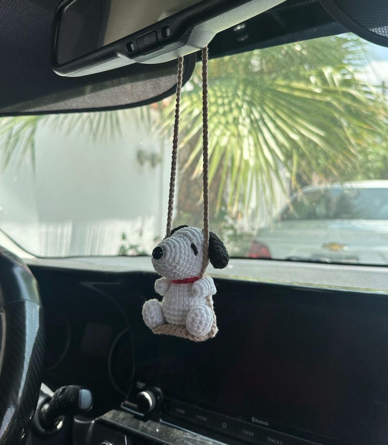 Puede incluir: Un adorno de coche de Snoopy hecho a mano con ganchillo. El adorno presenta un cuerpo blanco, orejas negras y un collar rojo. Est&aacute; suspendido por una cuerda marr&oacute;n y cuelga del espejo retrovisor de un coche. El fondo muestra una palmera y un interior de coche.
