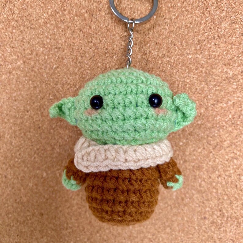 May include: A crocheted keychain featuring a green character with large ears, black eyes, and rosy cheeks. The character wears a tan scarf and a brown outfit. The keychain has a silver metal ring.