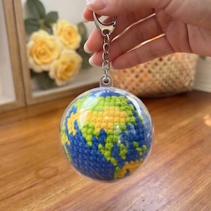 May include: A globe-shaped keychain with a clear plastic casing. The globe inside is crocheted with blue, green, and yellow yarn, depicting the continents and oceans. The keychain has a silver-colored clasp and chain.