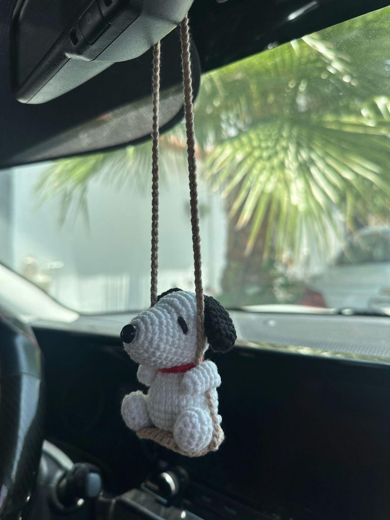 Puede incluir: Un adorno de coche de Snoopy tejido en blanco y negro con un collar rojo, colgado de cuerdas marrones. El adorno est&aacute; suspendido frente al interior de un coche, con un fondo borroso de palmeras.