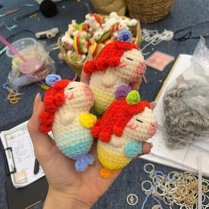 May include: Three handmade crochet mermaid dolls with red hair, smiling faces, and colourful tails. The dolls are held in a hand, with a basket of similar dolls in the background. The dolls are approximately 10 cm tall.
