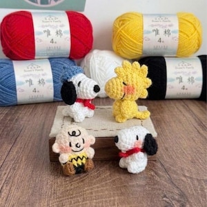 Könnte beinhalten: Handgefertigte Häkelfiguren von Snoopy, Woodstock und Charlie Brown, mit passenden Garnknäueln in Rot, Gelb, Blau, Schwarz und Weiß. Die Figuren werden auf einer Holzoberfläche präsentiert und zeigen die leuchtenden Farben und die detaillierte Handwerkskunst.