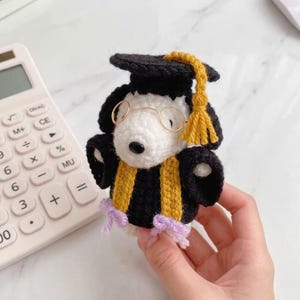 Op de afbeelding: Een gehaakte Snoopy-figuur in een afstudeeroutfit. De figuur is zwart en wit, met gouden details op de hoed en toga. Het heeft een kleine, ronde bril en een paarse strik. Een rekenmachine is zichtbaar op de achtergrond.